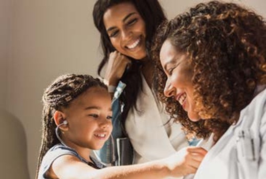 A pediatric patient listens to a physician's heartbeat through a stethoscope while her mother watches with a smile on her face.